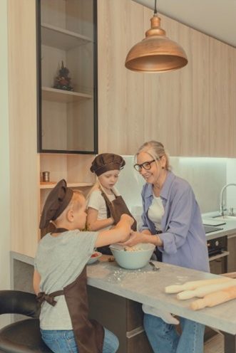 Grand mère avec ses deux petits enfants en train de faire des gateaux dans la nouvelle cuisine aménagée. Les meubles sont en bois clair. Il y a une vitrine en meuble haut dont l'encadrement est noir et qui laisse apparaitre 2 étagères à l'intérieur. Une suspension style industriel en cuivre est au dessus du plan de travail qui est en effet béton.