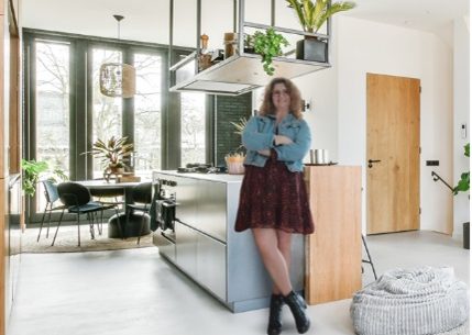 Linda Yakoub, décoratrice d'intérieur en Seine et Marne est adossée à un ilot de cuisine. L'ilot est en inox adossé à un meuble en bois clair. Au dessus se trouvé une structure en métal qui tient la hotte et des étagères tout ça suspendu au plafond.